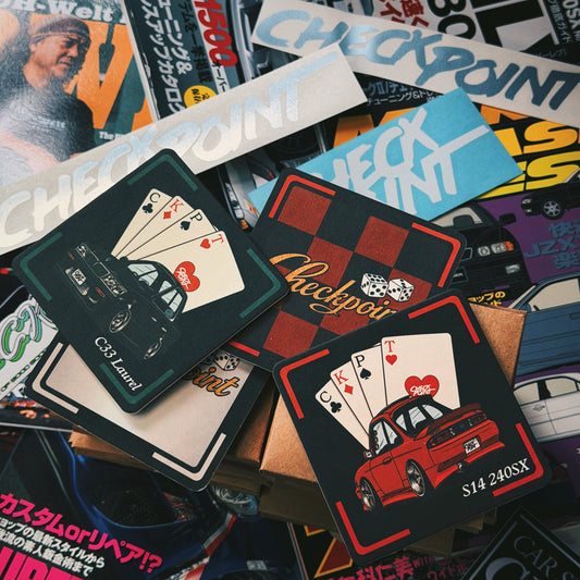 A pile of custom playing cards featuring car illustrations and Japanese text, layered over a background of magazine covers.