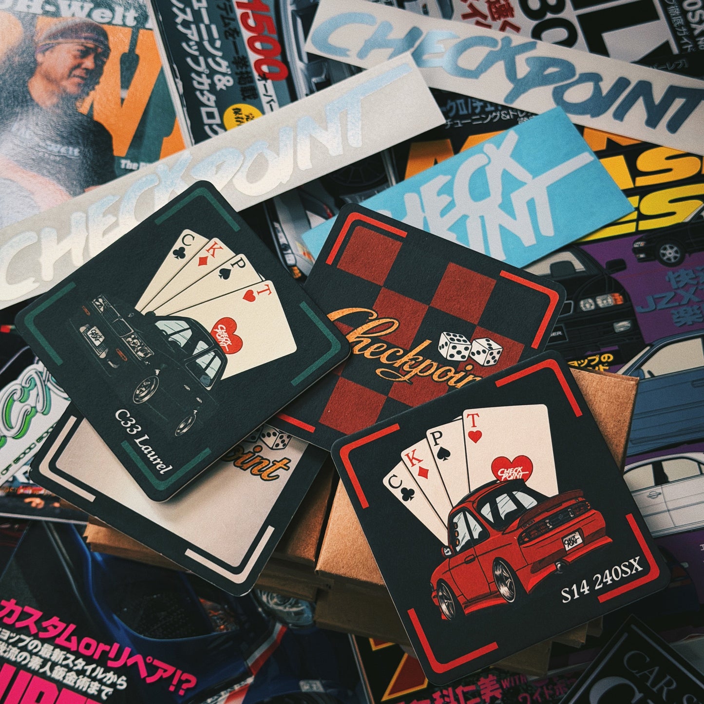 A pile of custom playing cards featuring car illustrations and Japanese text, layered over a background of magazine covers.