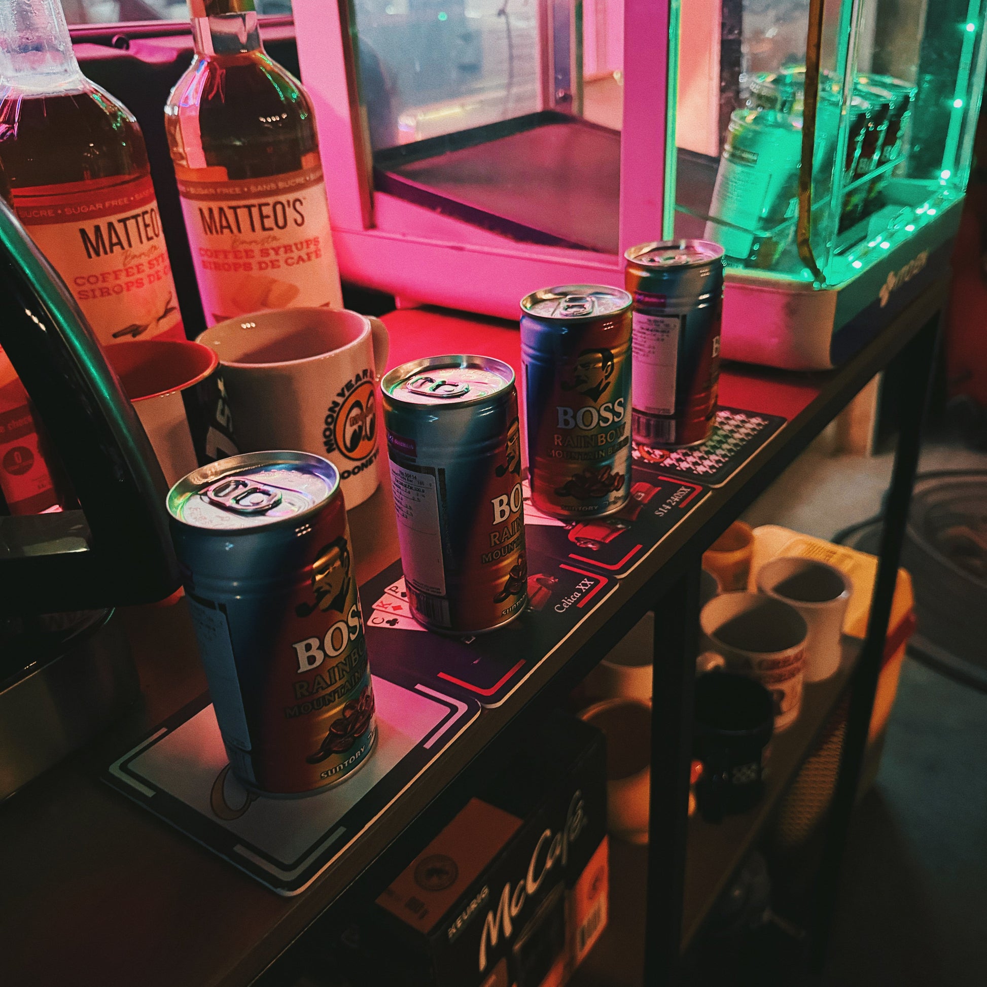 A bar counter scene with several beverage cans and bottles, including visible Mattéo's and Boss Hard liquor bottles, under pink and green neon lighting.