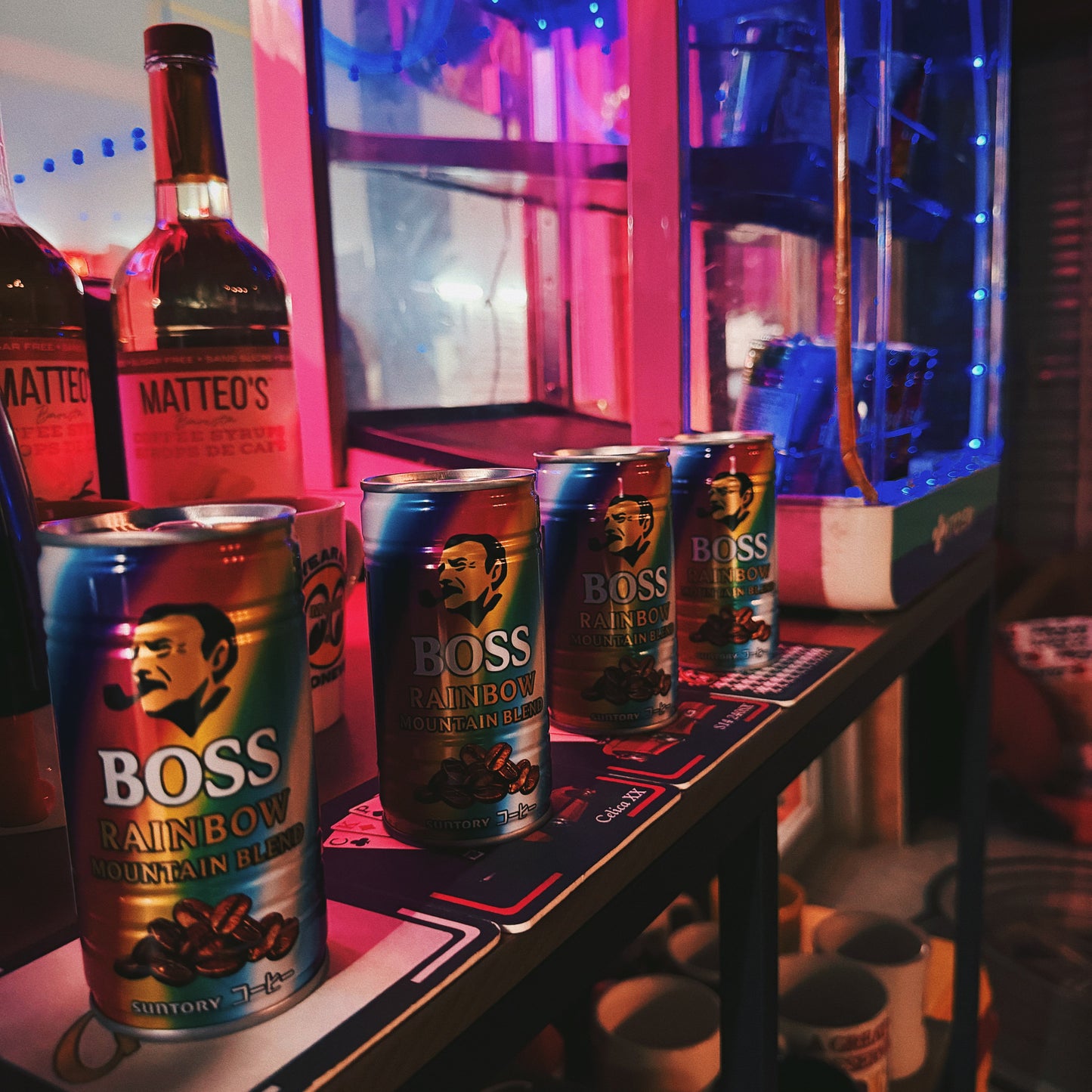 A row of canned coffee products, including Boss Rainbow Mountain Coffee and a bottle of Matteo's, displayed on a shelf in a dimly lit bar or cafe environment with pink and blue ambient lighting.